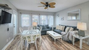 Ein Wohnzimmer mit Deckenventilator, einem an der Wand befestigten Fernseher, einem Esstisch mit Stühlen und einem Sofa mit Lampe.