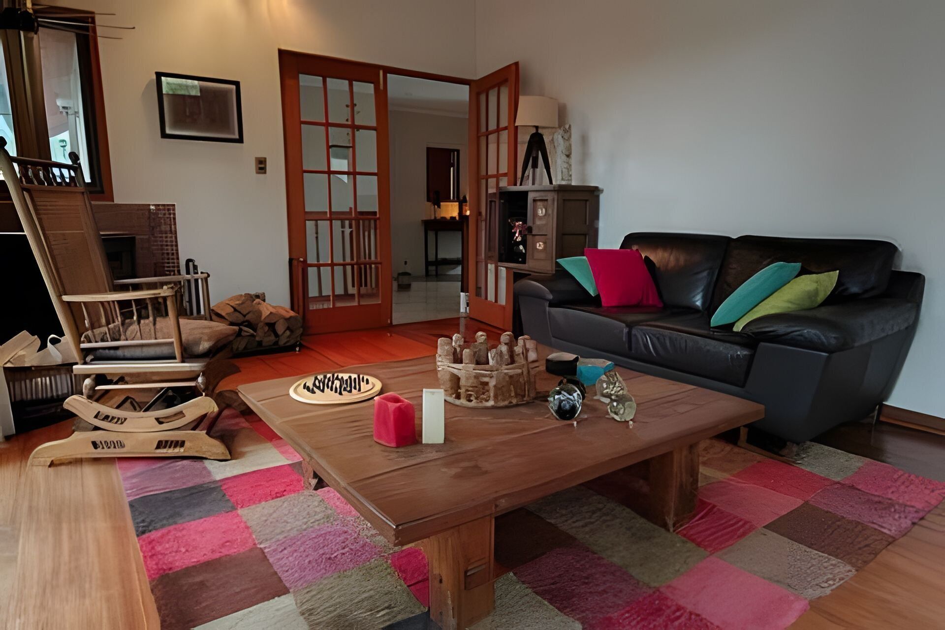 Una sala de estar con una mesa de centro de madera, un sofá de cuero negro con cojines coloridos, un sillón mecedor de madera y una puerta corredera tradicional japonesa.