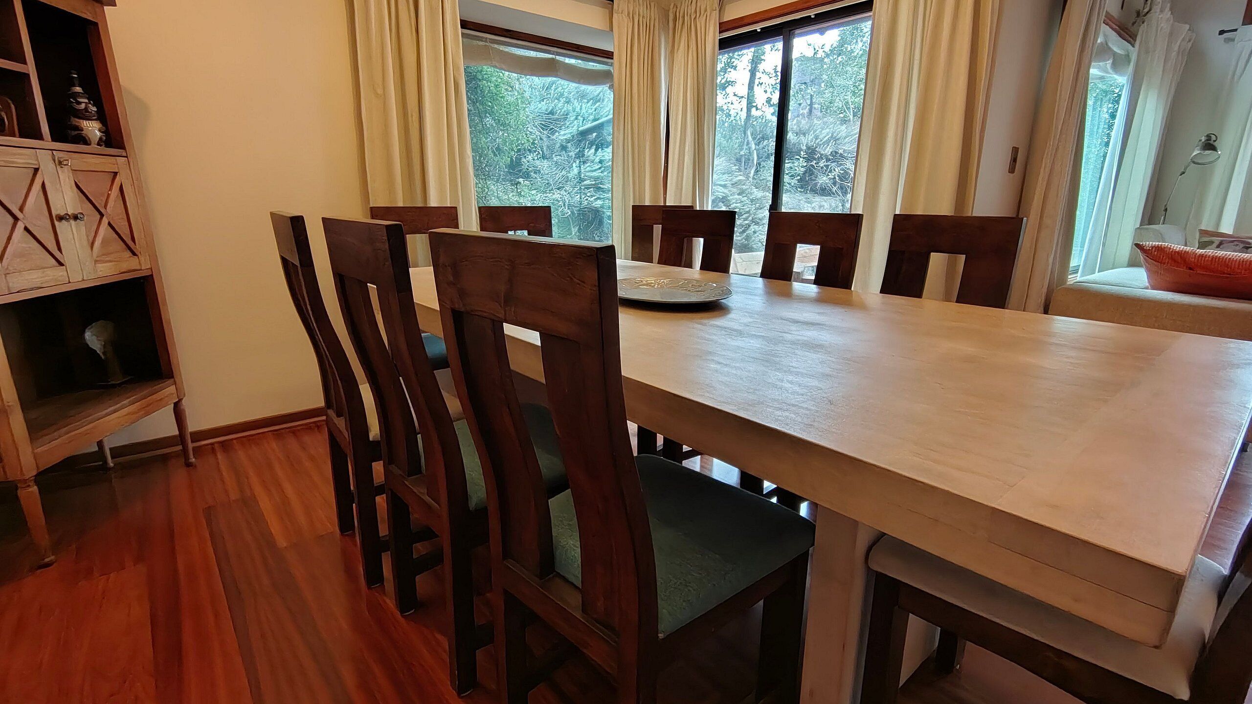 Uma área de refeições com uma mesa e cadeiras de madeira, uma janela com cortinas e uma estante com objetos decorativos.