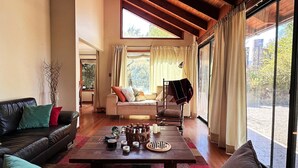 A cozy living room with a sofa, a coffee table with decorative items, a chair, and a view through large windows. - Oceana Suites - Laguna Aculeo (Paine)