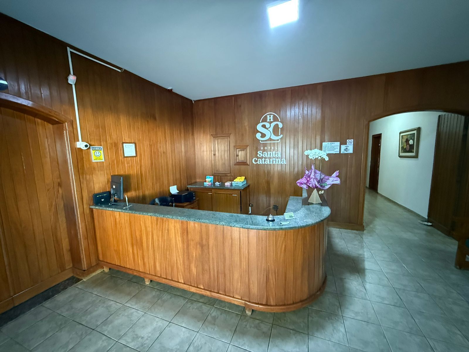 A hotel reception desk with a wooden finish, a logo above the desk, and a flower arrangement.