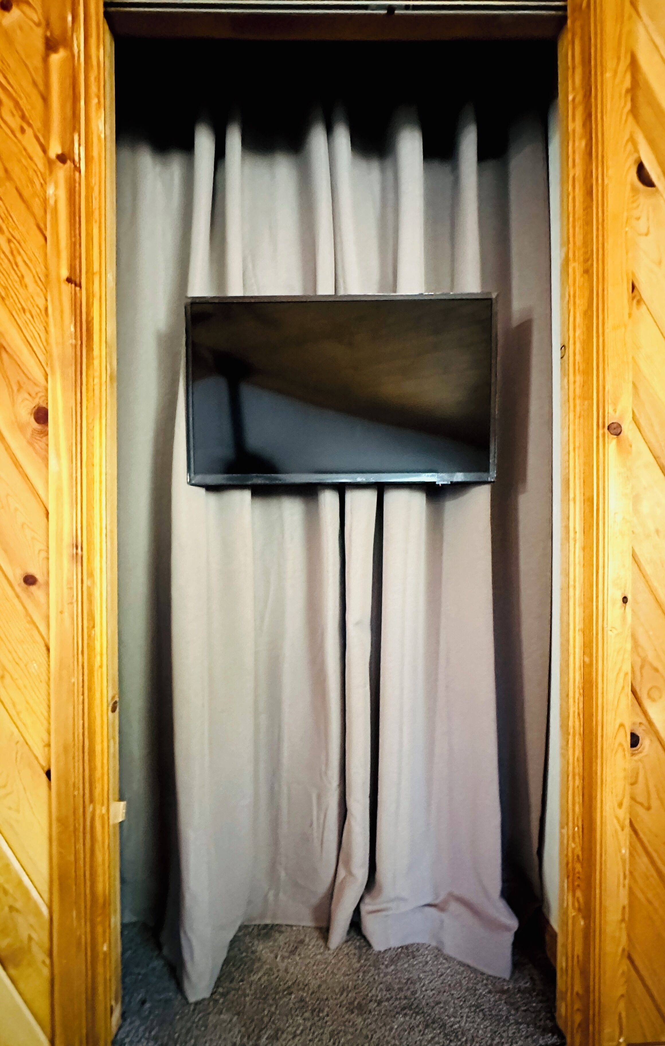 Un placard en bois avec un téléviseur à écran plat fixé au mur du fond, des rideaux blancs et un sol recouvert d’une moquette.