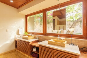 Une salle de bain avec deux lavabos, des meubles en bois, un miroir et une fenêtre donnant sur un paysage verdoyant.