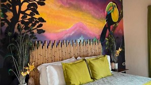 Un dormitorio con cama, cabecera de listones verticales, un mural colorido en la pared y una mesita de noche con un jarrón de flores.