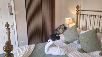 A neatly made bed with a green quilt, a white towel, and a swan-shaped towel arrangement. There is a lamp on a bedside table, a striped wall panel, and a door with a 'no smoking' sign.