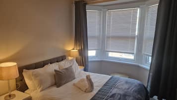 A neatly made bed with white and grey bedding, a bedside lamp, and a window with blinds.