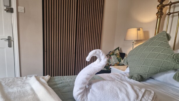 Ein Hotelzimmer mit Bett, einer grünen gesteppten Bettdecke, einem weißen, als Schwan geformten Handtuch, einem grünen Strickkissen, einer Lampe und einer Wandverkleidung aus Holzlamellen.