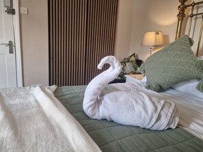 A hotel room with a bed, a green quilted bedspread, a white towel shaped like a swan, a green knitted pillow, a lamp, and a wooden slat wall panel.