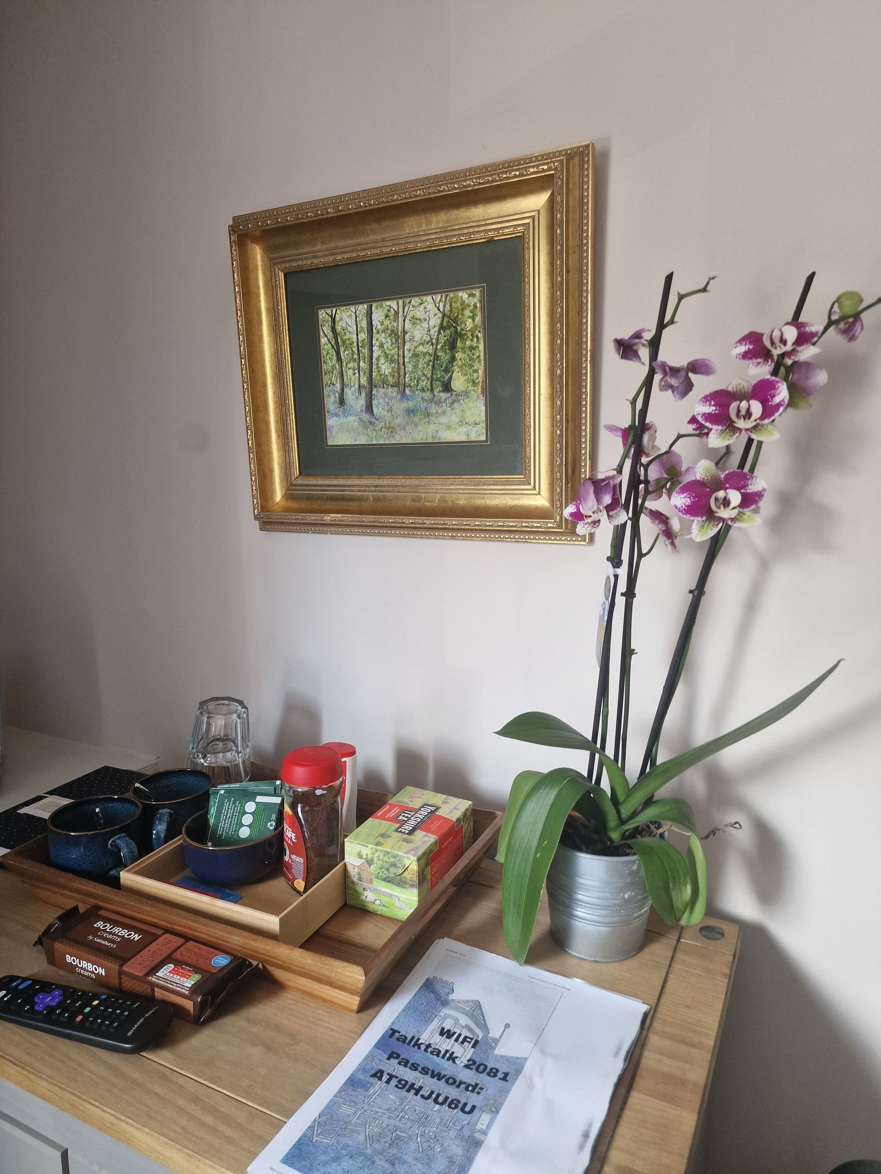 Een houten bureau met een lijstwerk schilderij, een potorkidee, en diverse voorwerpen, waaronder een afstandsbediening, potten en een doos bourbon.