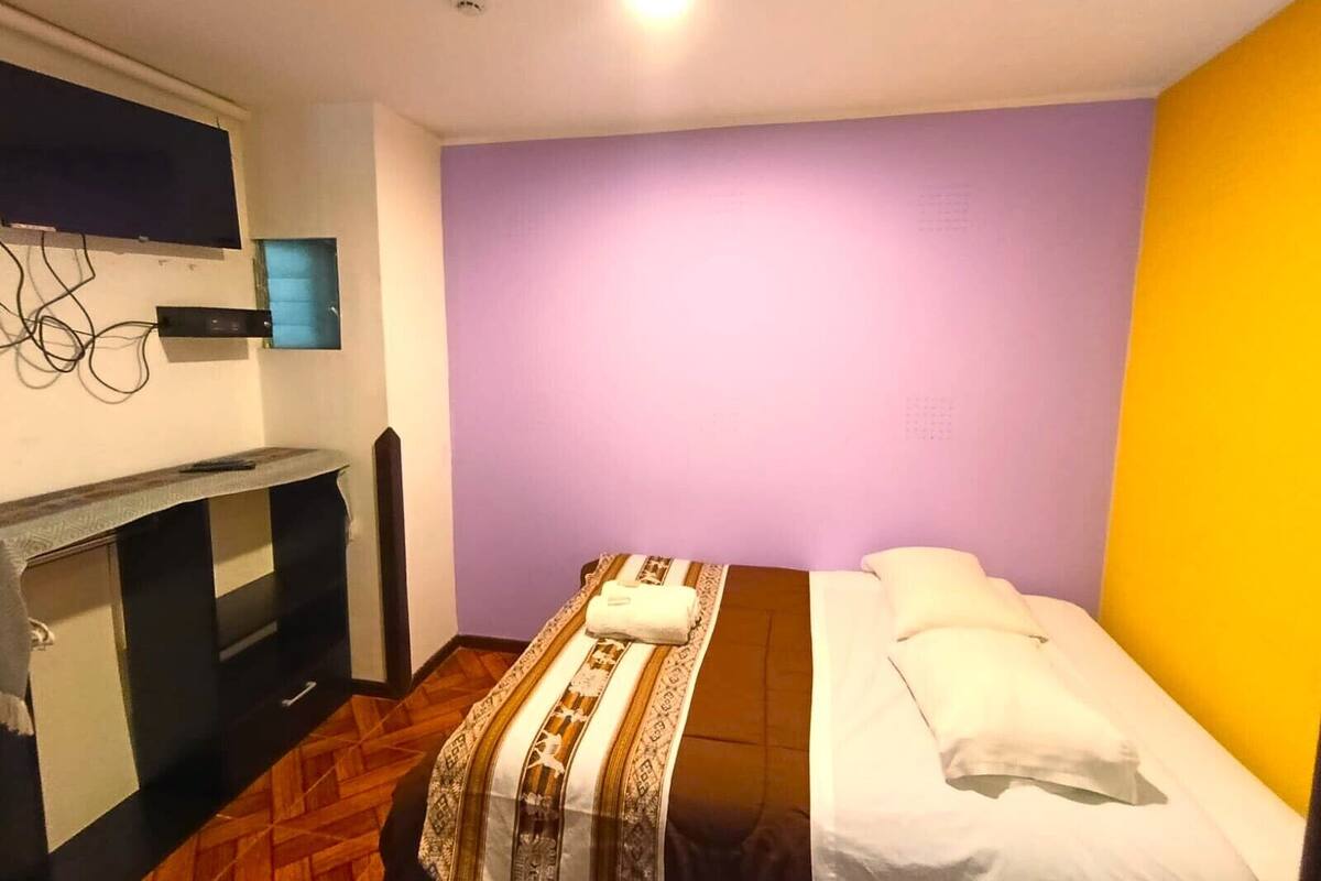 Une pièce comprenant un lit, un bureau, une chaise, un téléviseur fixé au mur et un luminaire au plafond.