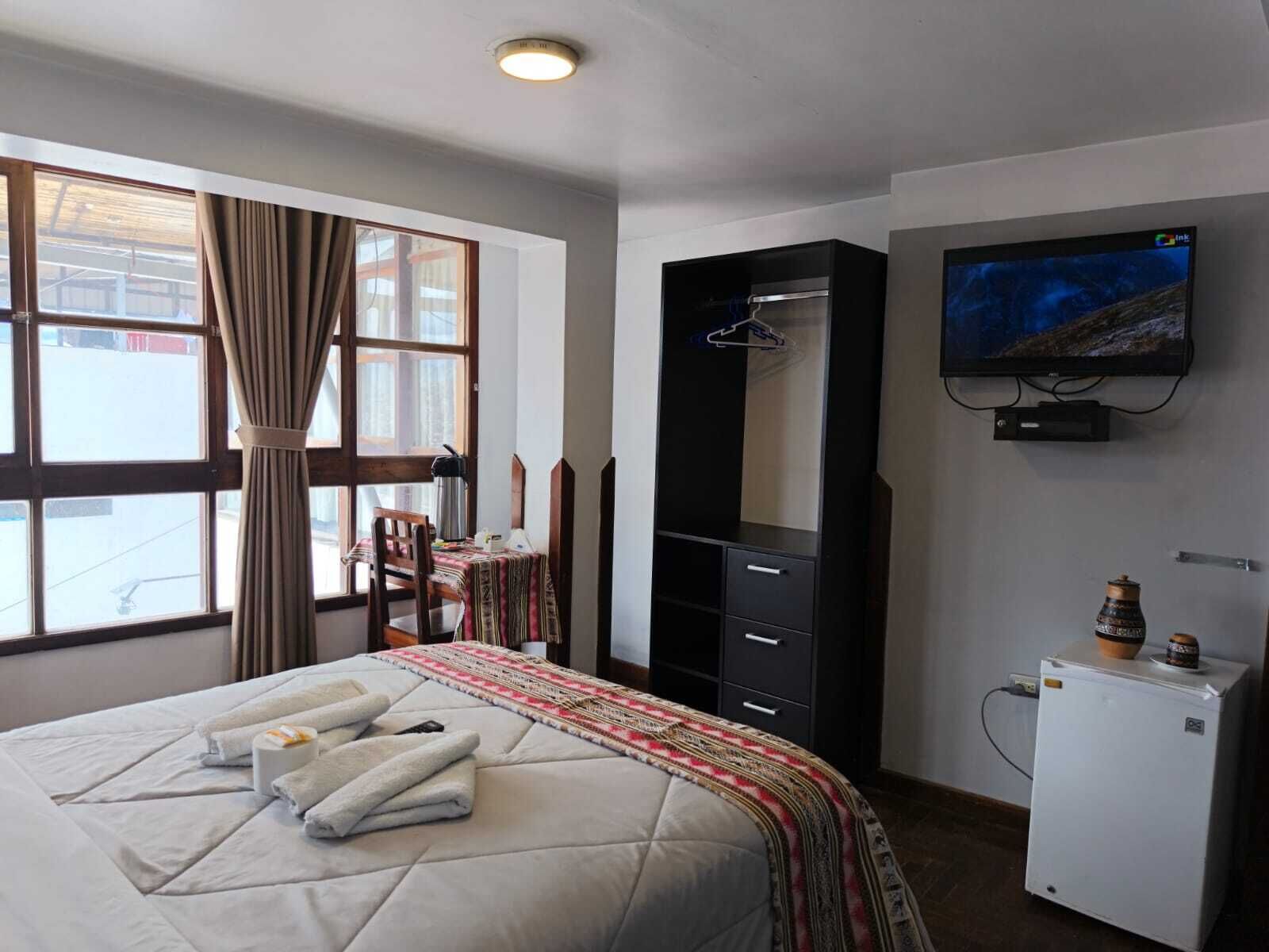 A hotel room with a bed, a television, a wardrobe, a desk with a chair, and a small refrigerator.