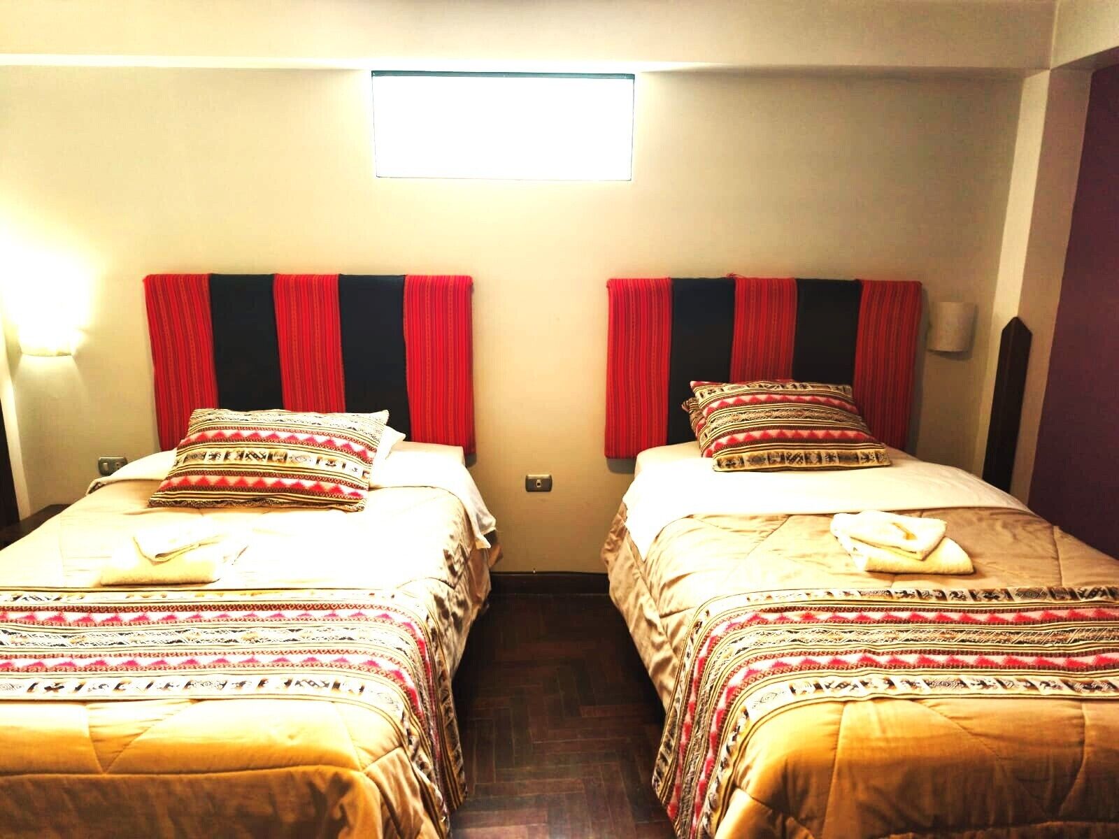 A room with two beds, each with patterned bedding and pillows, a small window above the beds, and a wall-mounted light fixture.