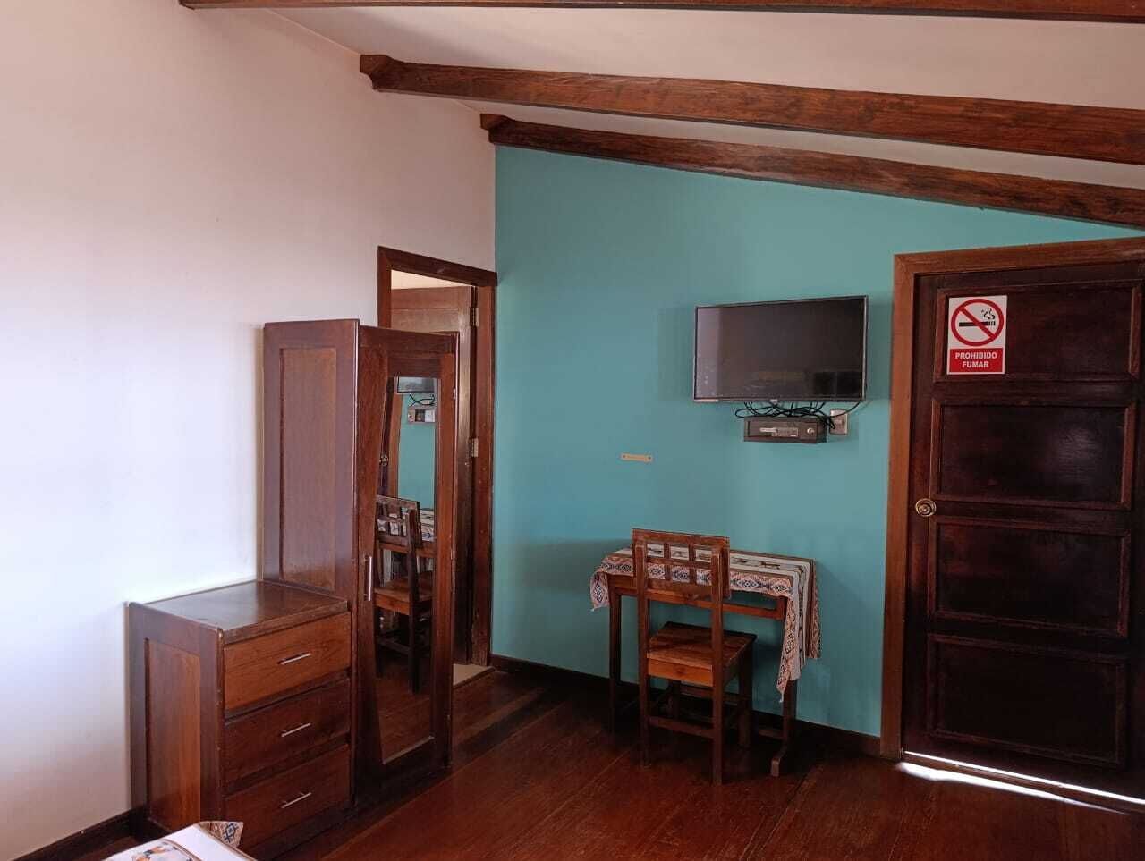 Quarto com guarda-roupa de madeira, uma pequena mesa com cadeiras, televisão na parede e porta com placa de proibido fumar.