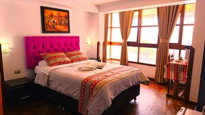 Een hotelkamer met een groot bed, een schilderij aan de muur, een raam met gordijnen, en een klein tafeltje met tafelkleed.
