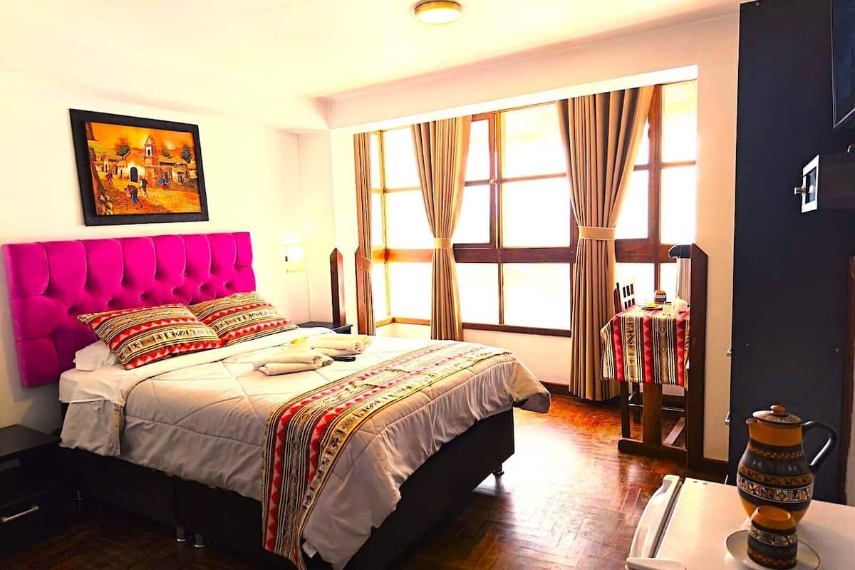 Une chambre d’hôtel avec un lit, un tableau au mur, une fenêtre avec des rideaux, une petite table avec une théière et une porte.