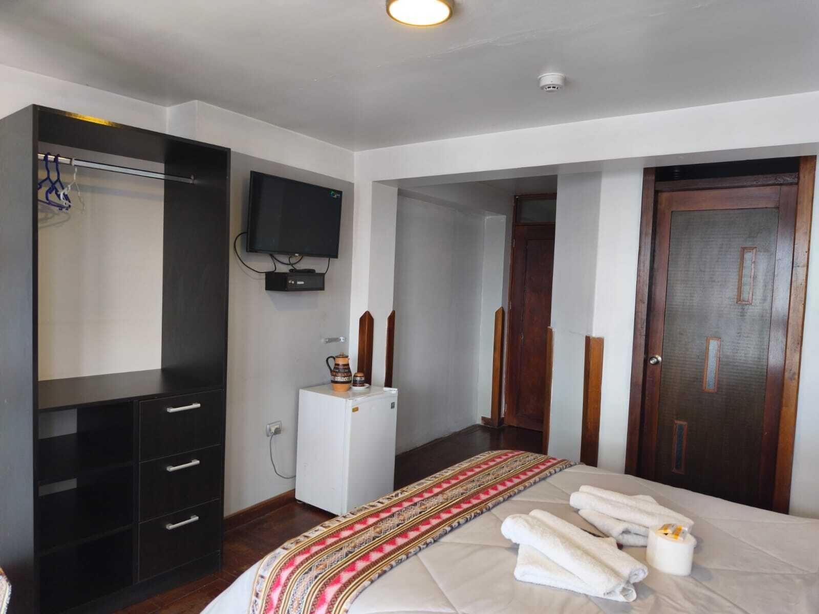 A hotel room with a bed, a TV, a small fridge, a wooden door, and a wardrobe.