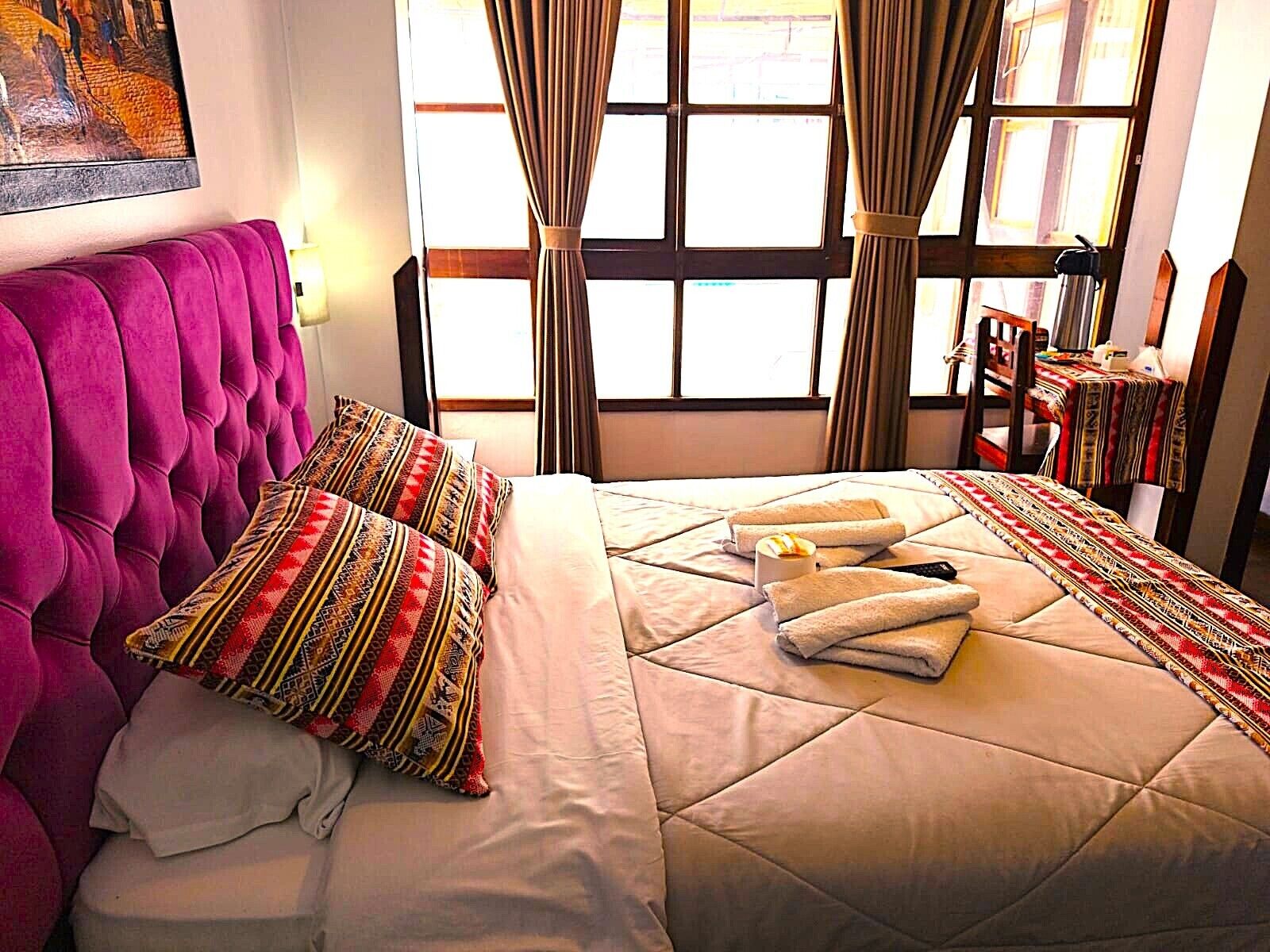 A hotel room with a tufted headboard, patterned pillows, a bed with a quilt, and a window with curtains.