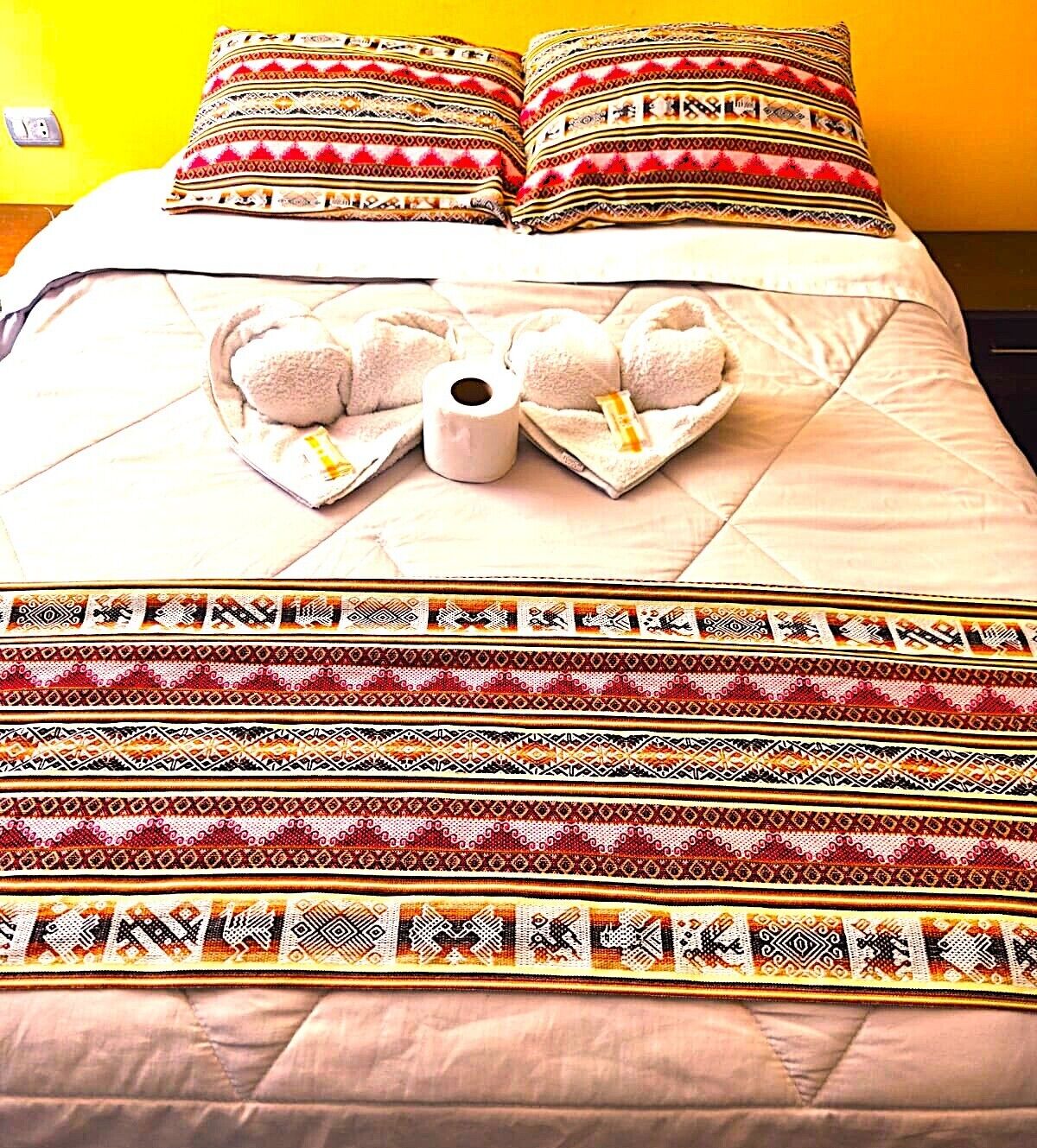 A bed with a patterned bedspread, pillows with a similar design, and a heart-shaped arrangement of pillows with a roll of toilet paper and a bottle.