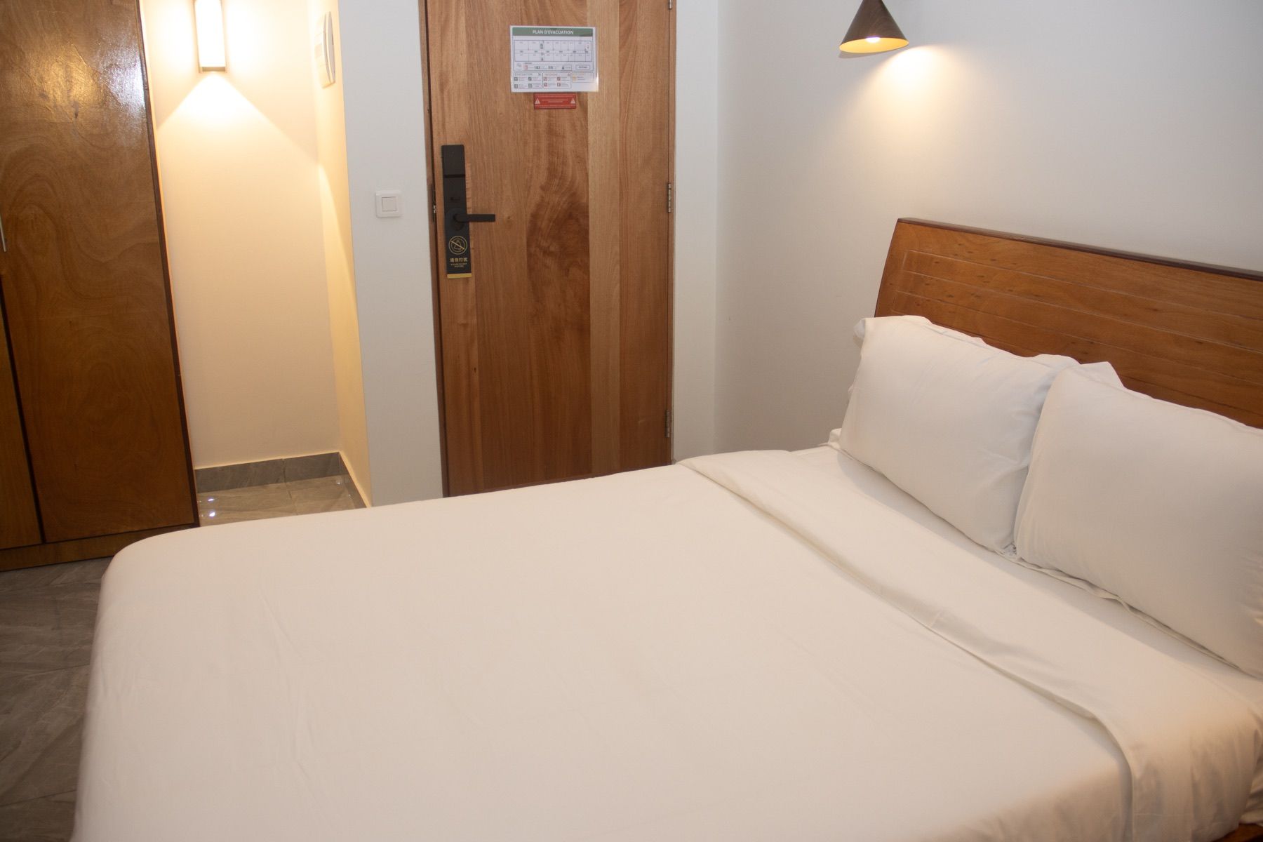 A hotel room with a wooden door, a bed with white bedding, and a wall-mounted light fixture.