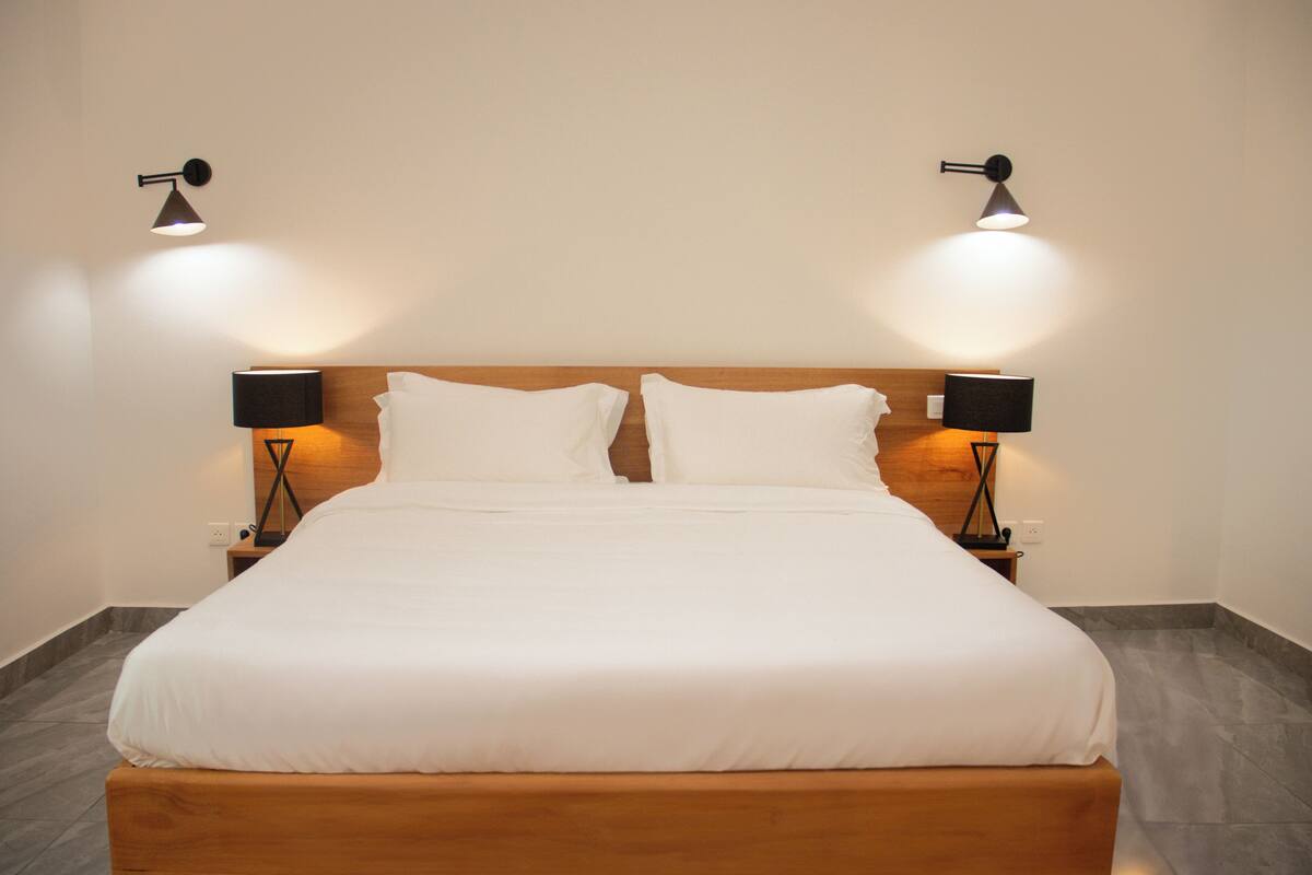 A neatly made bed with white linens, flanked by two bedside tables with lamps, in a room with a plain wall and a tiled floor.