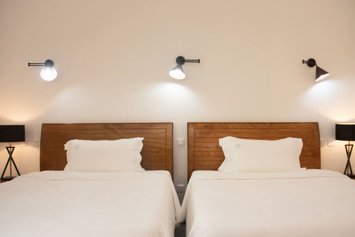 A hotel room with two beds, wooden headboards, and three wall-mounted lamps.