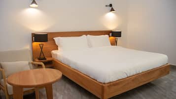 Quarto individual com uma cama e cabeceira de madeira, duas luminárias de mesa, uma mesinha de madeira e uma cadeira branca.