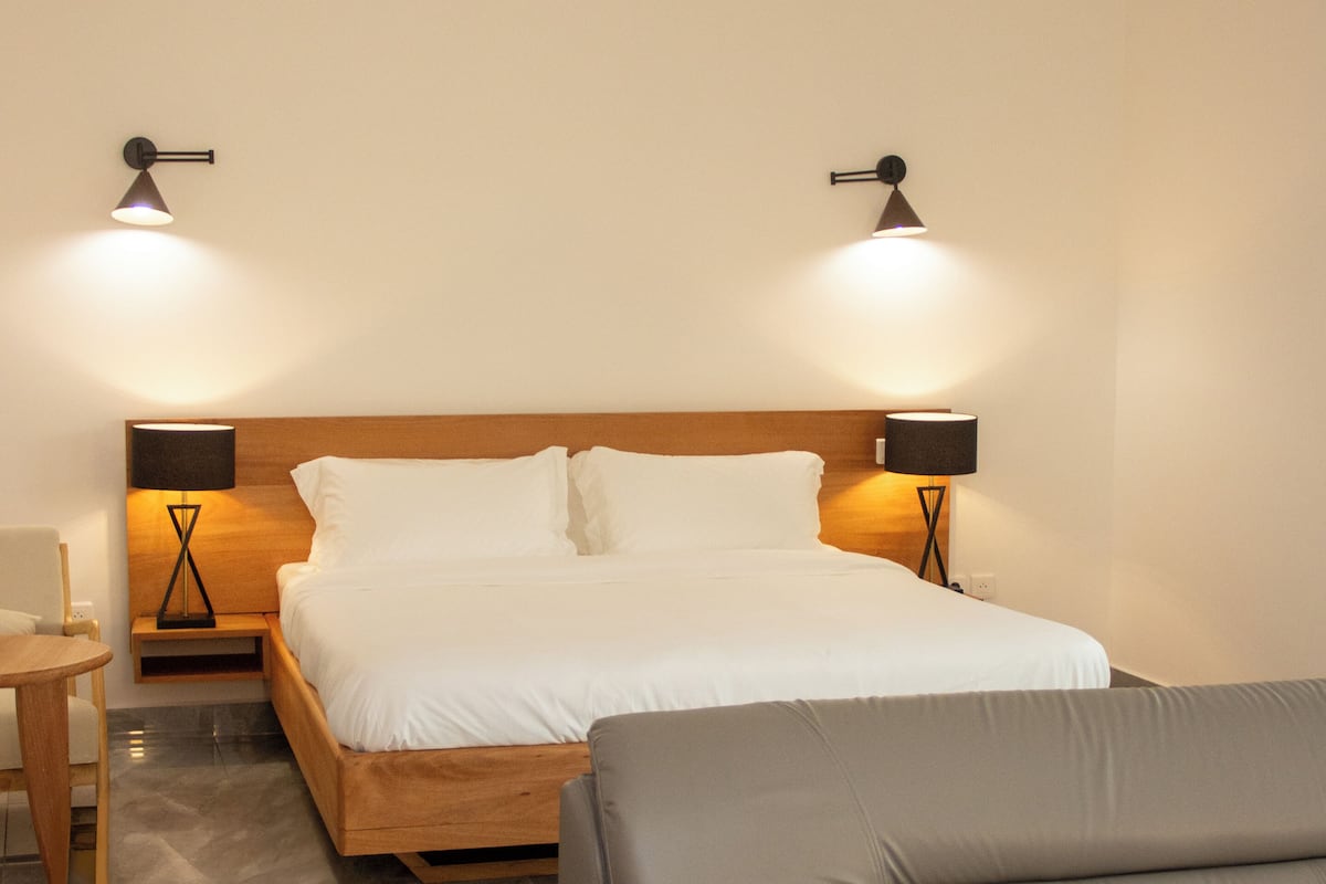 A hotel room with a bed, two bedside lamps, a wooden headboard, a grey sofa, and a small table.