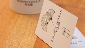 Una tazza bianca con un motivo a foglia e testo, e un biglietto da visita con lo stesso design e testo su una superficie in legno.
