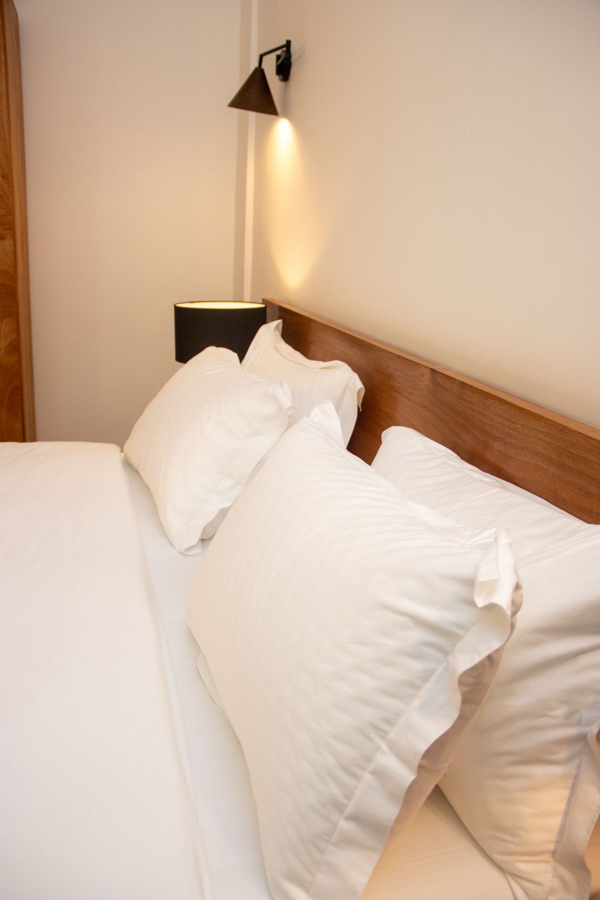 Un lit bien fait, avec du linge de lit blanc et des oreillers, une tête de lit en bois et une lampe fixée au mur.