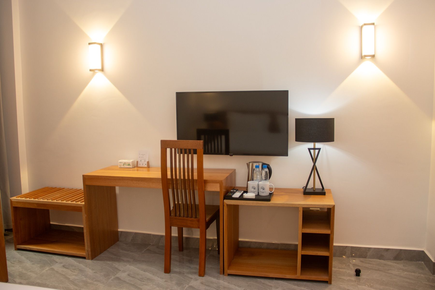 A room with a wooden desk, a chair, a flat-screen TV, a lamp, and a small table with a coffee maker and cups.