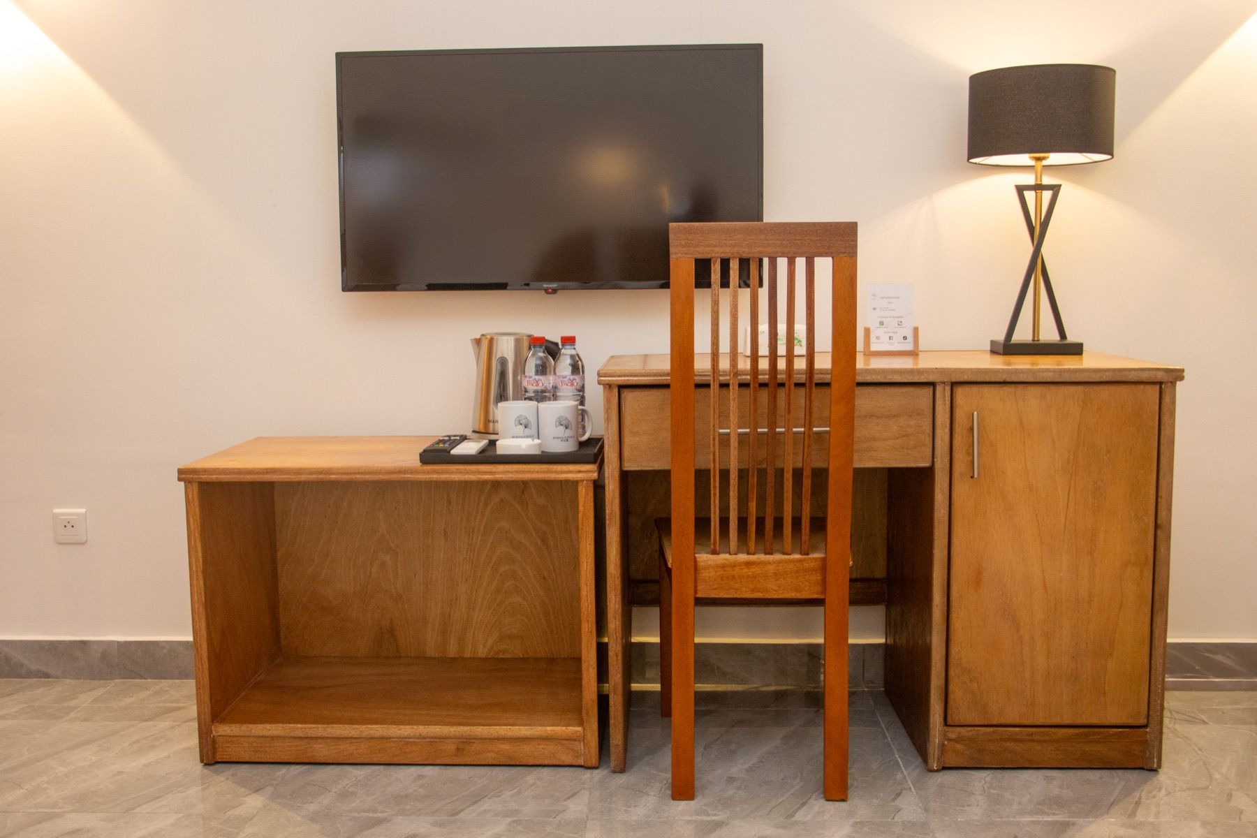 Ein kleines Hotelzimmer mit Schreibtisch aus Holz, einem Sessel, einem an der Wand montierten Fernseher und einer Stehlampe.