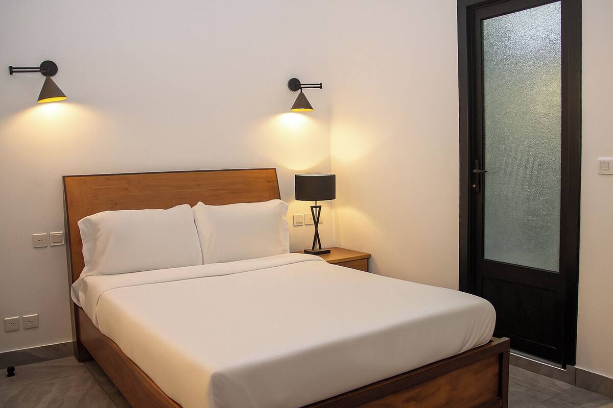 A neatly made bed with white linens, a wooden headboard, and two wall-mounted lamps on either side.