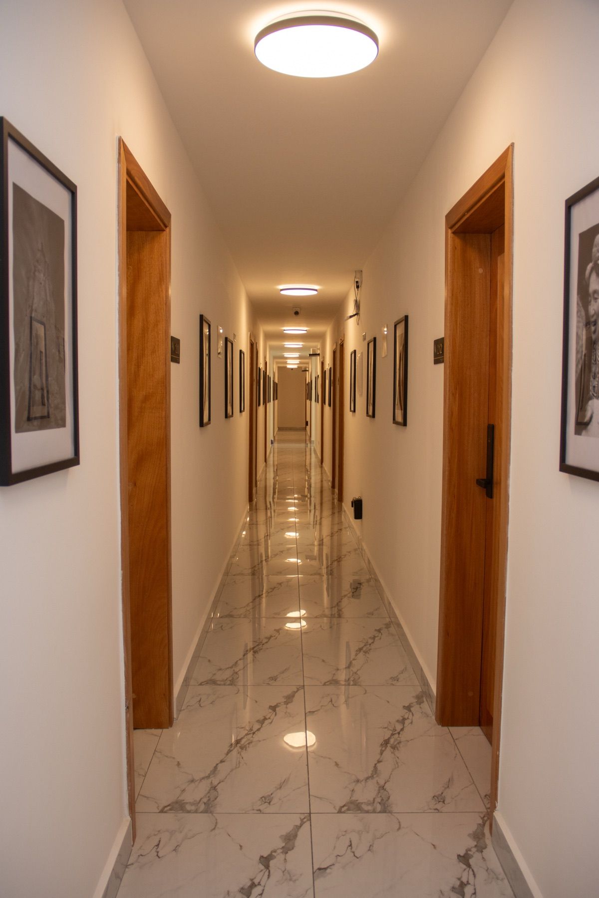 Corredor de hotel com portas de madeira, quadros nas paredes e piso de mármore.