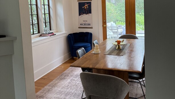 Un coin repas avec une table en bois, des chaises et un fauteuil bleu. Un poster est accroché au mur, et une fenêtre offre une vue sur la verdure.