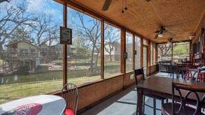 A covered outdoor dining area with large windows overlooking a lake and buildings. - Lakeside Rock house on Lake LBJ! (Llano County)
