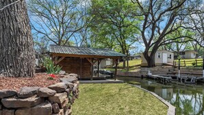 A rustic cabin with a stone wall and a pond, surrounded by trees and a grassy lawn. - Lakeside Rock house on Lake LBJ! (Llano County)