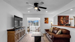 Una sala moderna con televisor de pantalla plana, ventilador de techo, mueble de madera, sofá de cuero marrón y comedor con mesa y sillas.