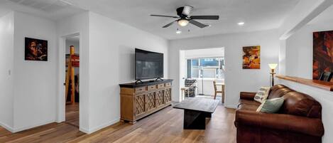 Ein geräumiges Wohnzimmer mit Holzboden, einer Ledersofa, einem Fernseher auf einer Holzkommode und einem Deckenventilator.