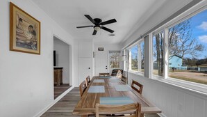 Un comedor con una larga mesa de madera, sillas, un ventilador de techo y ventanas amplias que dan vista al exterior.
