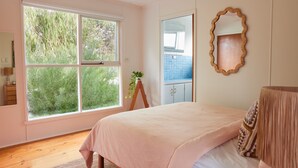 A bedroom with a large window, a wooden dresser, a bed with a light-colored bedspread, and a mirror on the wall. - Lohr Ave Holiday Home (Inverloch)