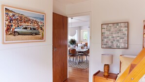 A room with a wooden door, a framed beach photo, a dining area with a table and chairs, and a lamp on a side table. - Lohr Ave Holiday Home (Inverloch)