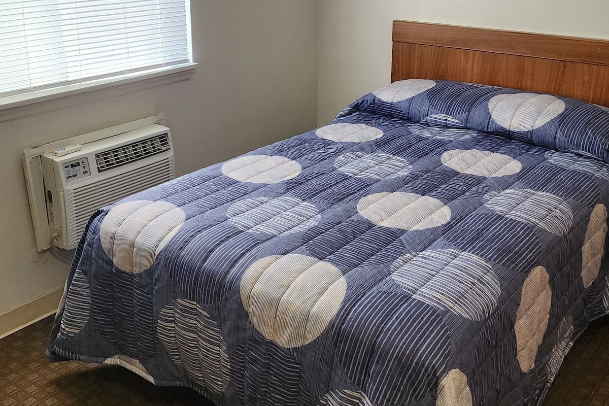 Une chambre à coucher avec un lit, une fenêtre avec des stores, un appareil de climatisation et un sol recouvert de moquette.