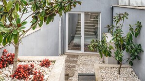 Un bâtiment moderne à l’extérieur gris, avec un escalier menant à une porte en verre, et un espace jardin agrémenté de galets blancs et de feuillage rouge.