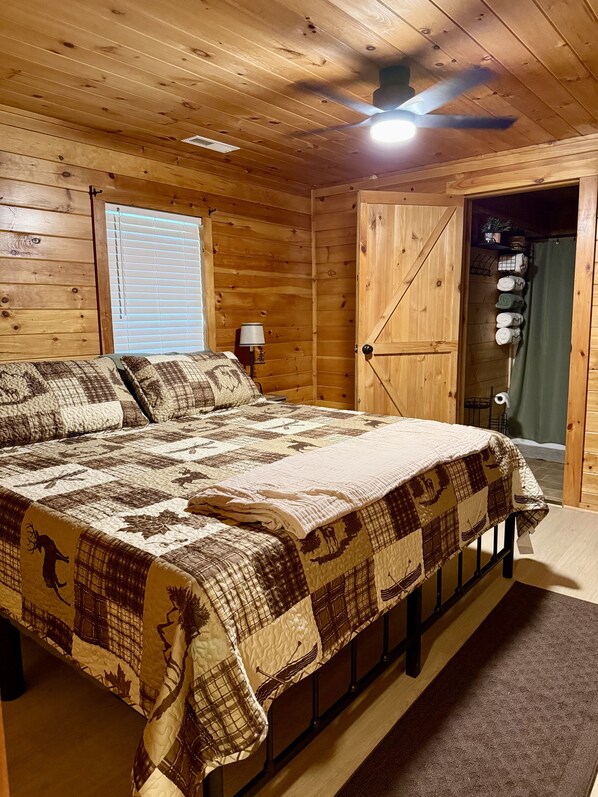Een slaapkamer in een houten chalet, met een bed, een plafondventilator, een raam met jaloezieën en een deur naar een andere kamer.