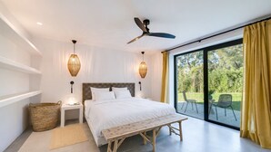 Une chambre moderne avec un grand lit, des tables de chevet, un ventilateur de plafond et une vue sur un jardin, accessible par une porte-fenêtre coulissante.