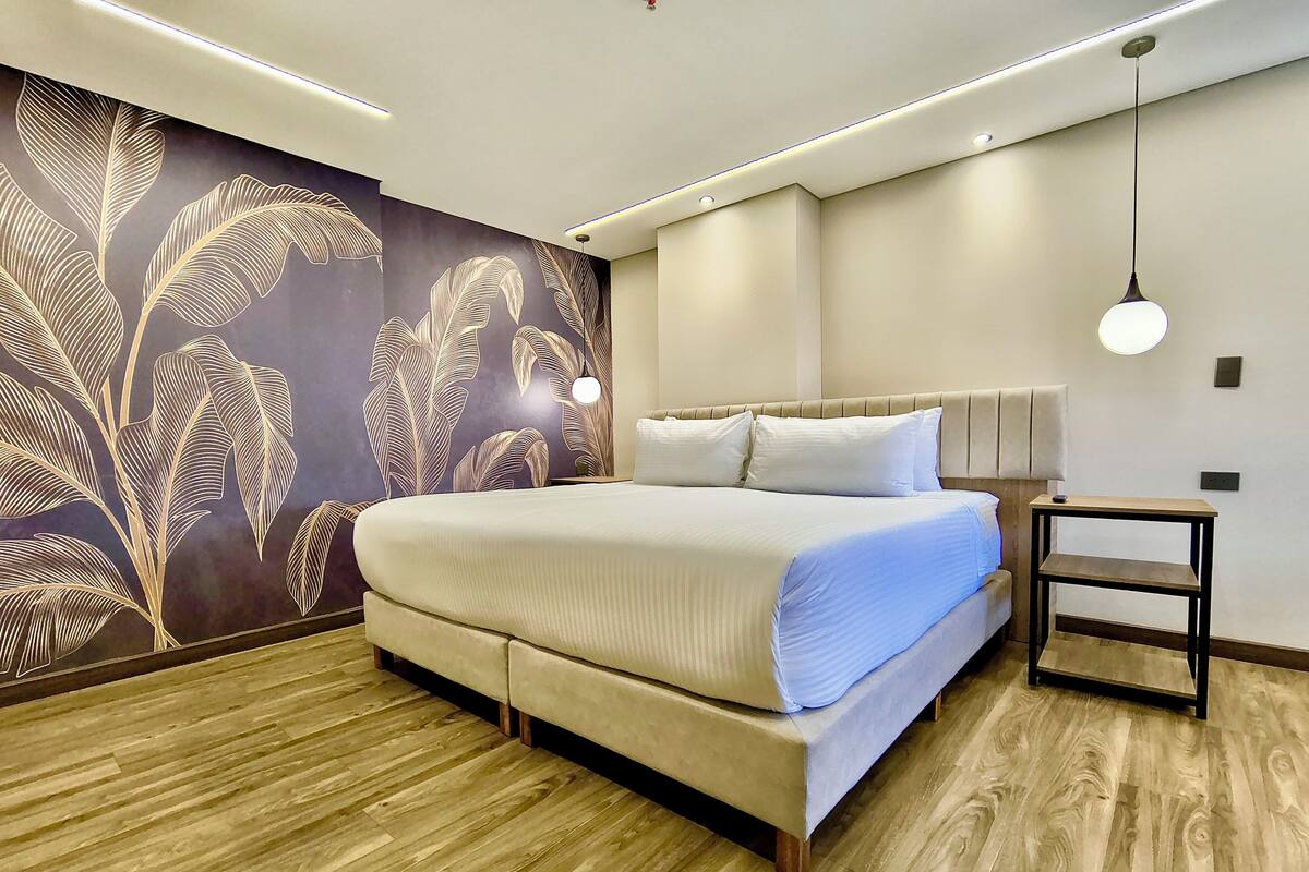 Un dormitorio moderno con una cama grande, suelo de madera, pared con diseño de hojas y una mesita de noche con lámpara.