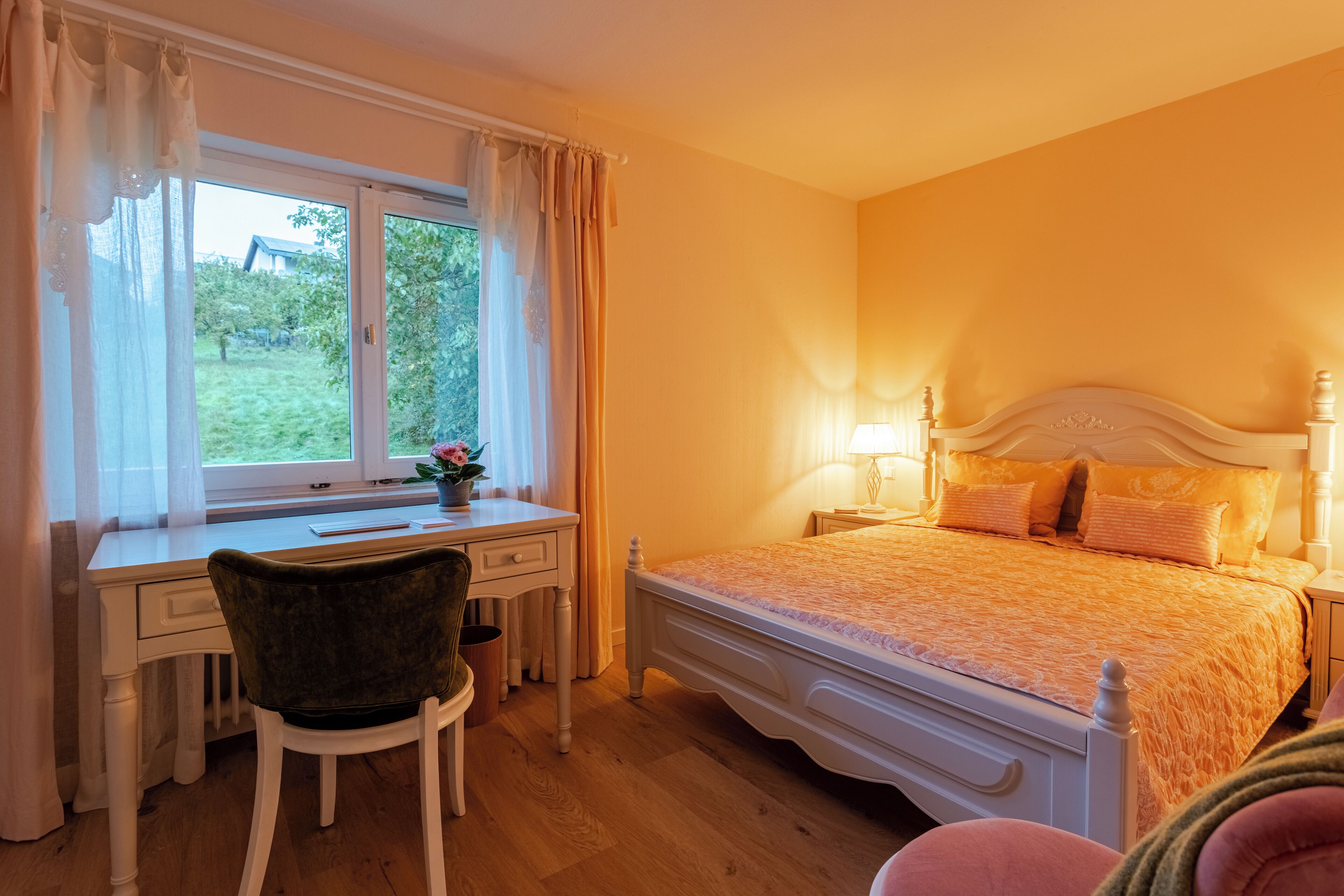 A bedroom with a bed, bedside table, lamp, chair, desk, and window with curtains.