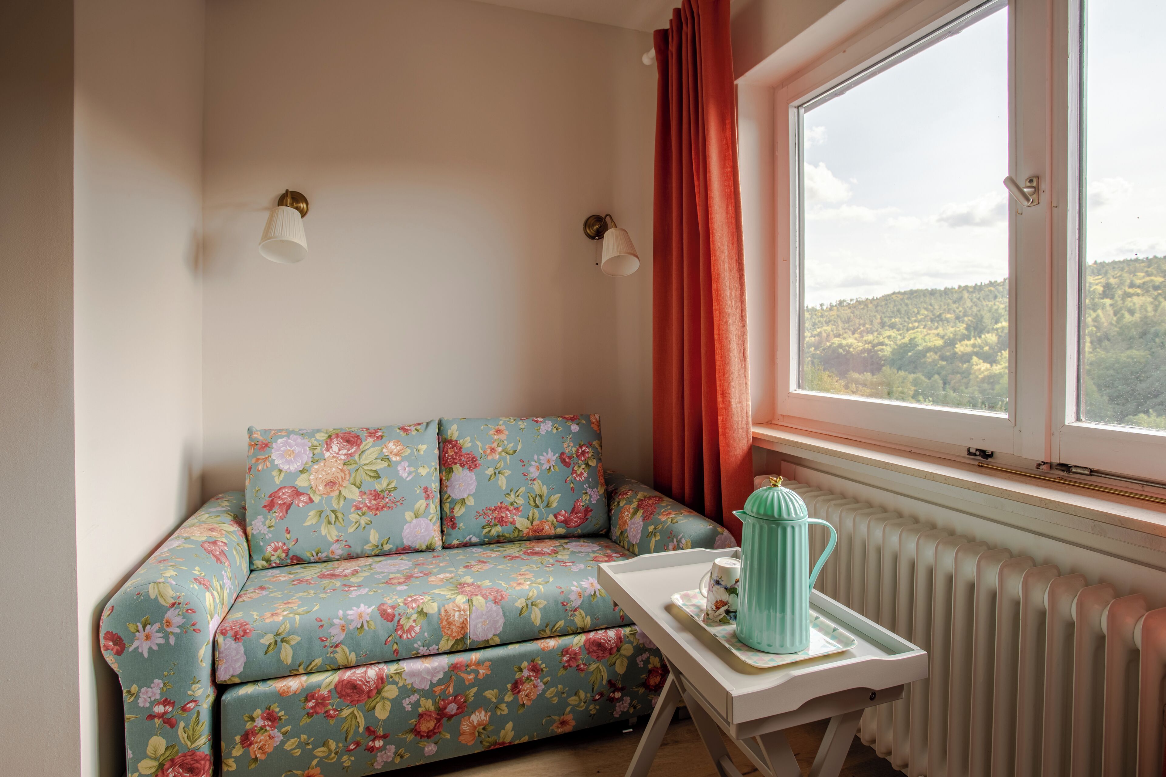 Et hyggeligt rum med en blomstret sofa, et lille bord med et te-service, en radiator og et vindue med gardiner.