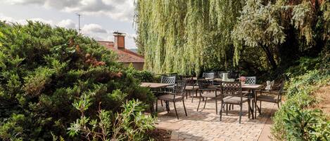 An outdoor patio with brick flooring, surrounded by dense greenery and a weeping willow tree.