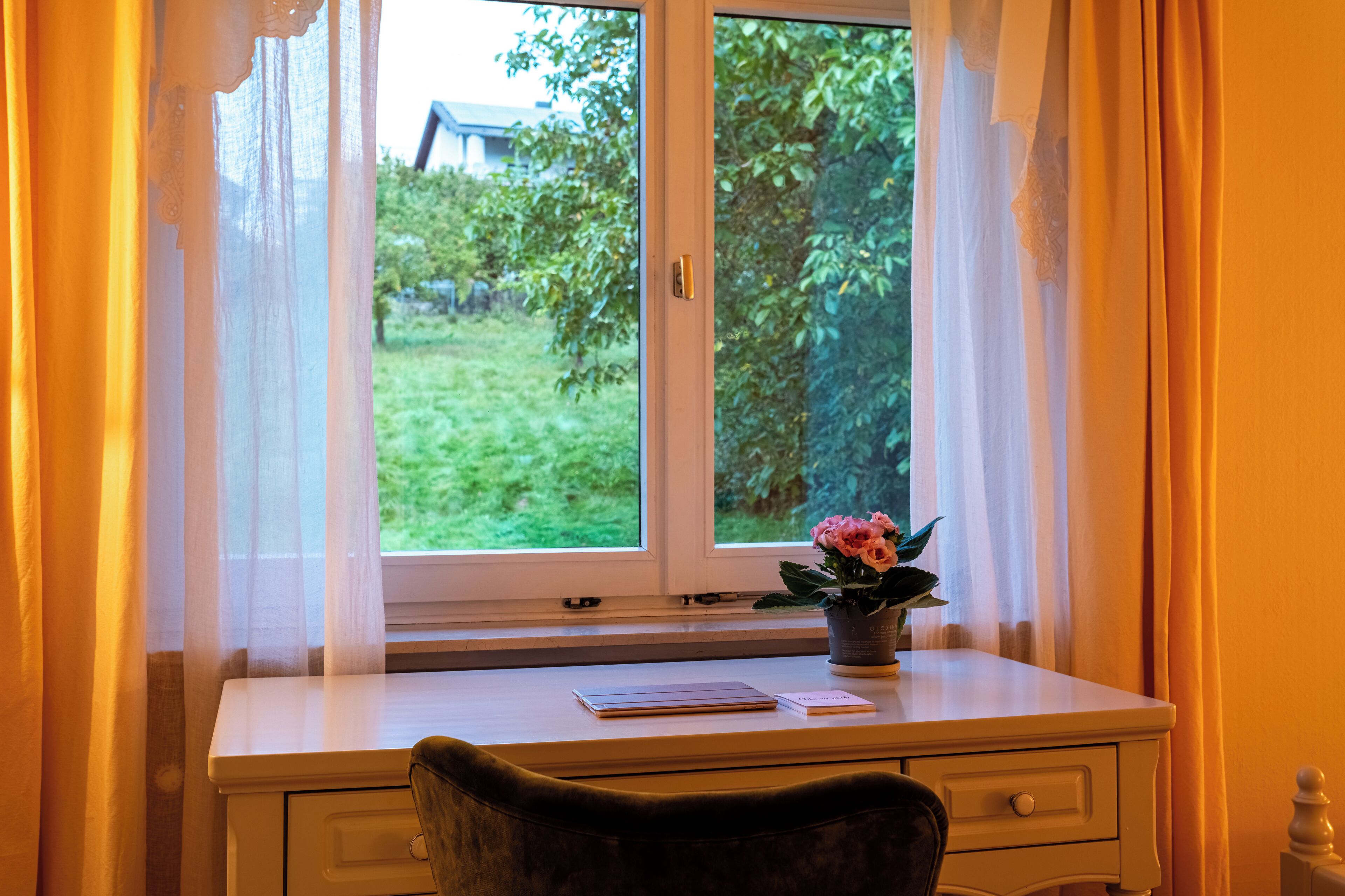 Quarto com escrivaninha e cadeira, janela com cortinas e uma planta em vaso no parapeito.